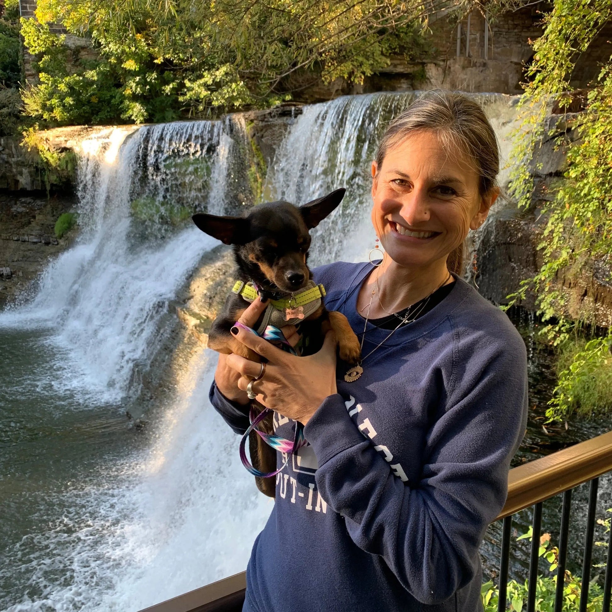A smiling woman in a blue sweatshirt holds a freshly pampered small black and tan dog, cleaned with Your Only Earths Happy Coat non-toxic dog soap. They stand on a wooden deck with a scenic waterfall and lush greenery. The sun casts a warm glow over the scene.