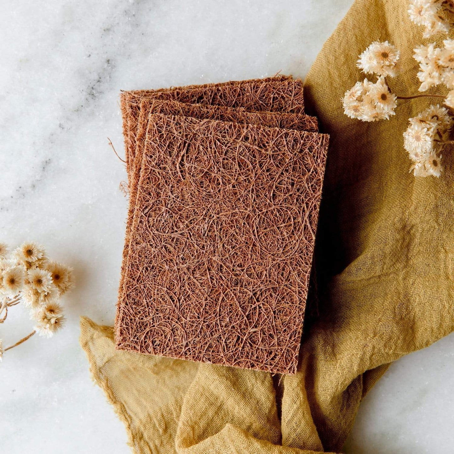 Two Coconut Kitchen Scrubbers from ZeroWasteStore.com, made of brown coconut fibers, are stacked on a light marble surface with dried flowers and a mustard yellow cloth, adding a rustic, natural touch to your kitchen.