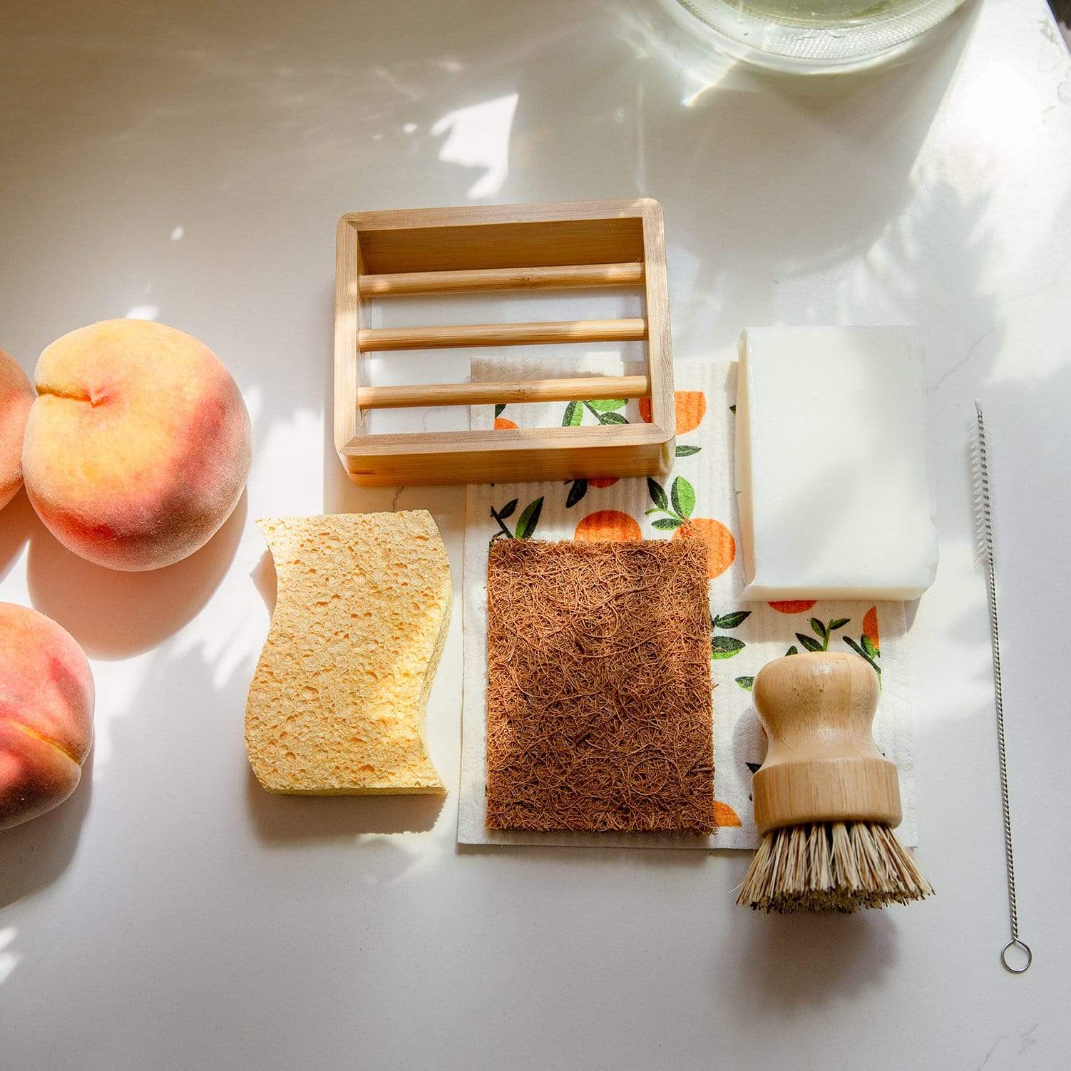 A flat lay of eco-friendly cleaning items: two peaches, a wooden soap dish, Cellulose Kitchen Sponges by ZeroWasteStore.com, coconut scrub pad, bar soap, wooden-handled brush, orange-patterned cloth, and a thin cleaning brush on a white surface.