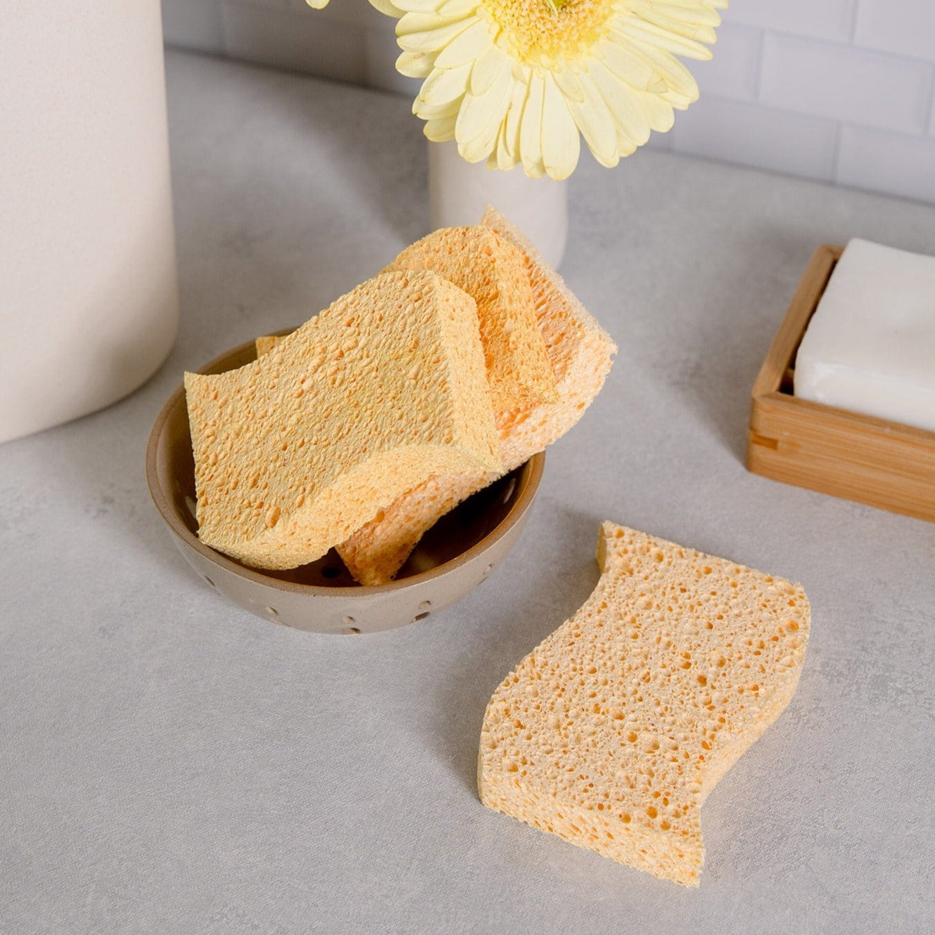 A small bowl contains several yellow Cellulose Kitchen Sponges from ZeroWasteStore.com on a light countertop. One sponge rests beside the bowl, with a yellow flower and a soap dish visible in the background.