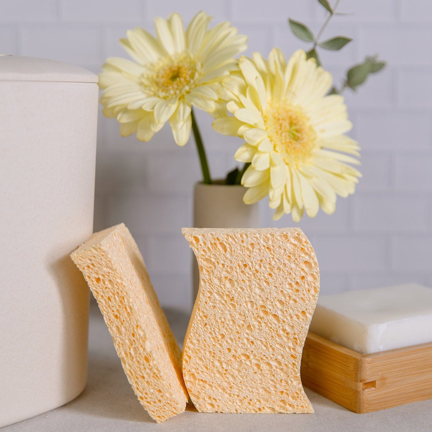 Two yellow Cellulose Kitchen Sponges from ZeroWasteStore.com stand upright beside a bar of soap in a wooden dish, with two pale yellow flowers in a vase and a white container in the background.