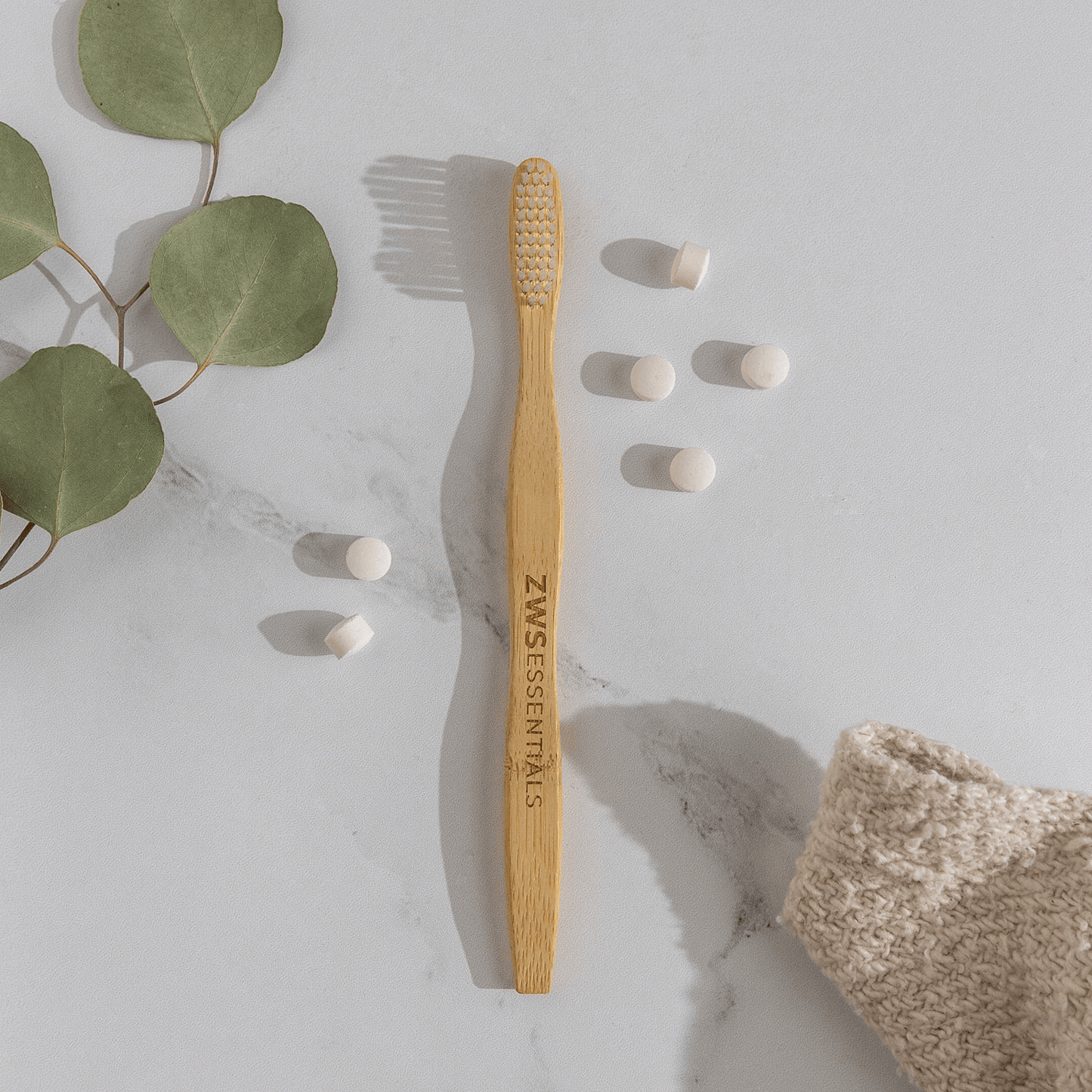 A ZeroWasteStore.com Bamboo Toothbrush rests on a white surface beside six toothpaste tablets, eucalyptus leaves, and a beige cloth—showcasing a zero waste toothbrush for your plastic free routine.
