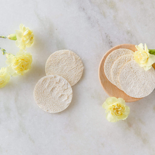 ZeroWasteStore.com Loofah Facial Disc Pack—exfoliating, round natural loofah sponges—rest on a wooden plate and marble surface with yellow carnations; perfect for facial exfoliation and fully compostable.