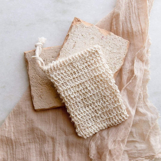 The Soap Saver Bag by ZeroWasteStore.com, crocheted from natural agave fiber with a drawstring, sits atop two bars of natural soap on crumpled fabric and marble—perfect for an eco-friendly, zero waste bathroom routine.