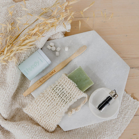 A flat lay of zero waste bathroom essentials, featuring a bamboo toothbrush, toothpaste tablets, a green bar of soap in the ZeroWasteStore.com Soap Saver Bag, a small glass vial, and dried wheat on a white hexagonal tray.
