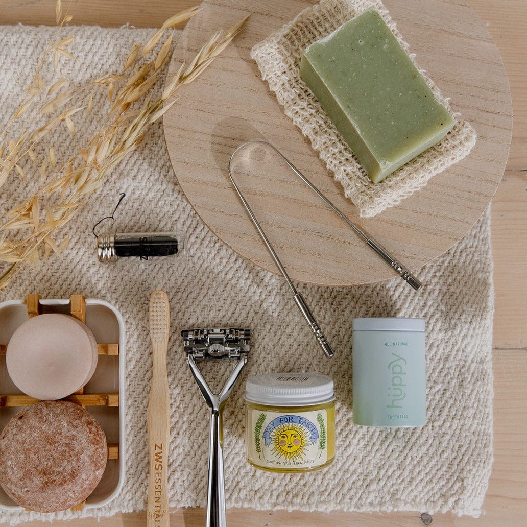 A flat lay of eco-friendly toiletries on a textured mat, including two round soaps, a green bar soap in the ZeroWasteStore.com Soap Saver Bag made from natural agave fiber, plus a toothbrush, safety razor, jar, deodorant stick, tongue cleaners, and black pod.