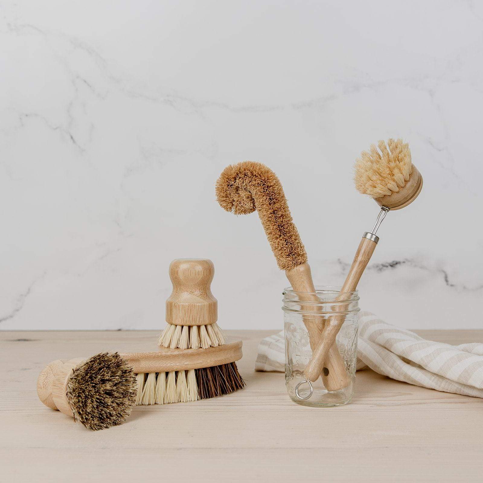 The Bottle Cleaning Brush by ZeroWasteStore.com, made of wood and natural bristles, sits arranged with other eco-friendly brushes on a light wood surface. Some are in a glass jar beside a striped cloth and marble background.