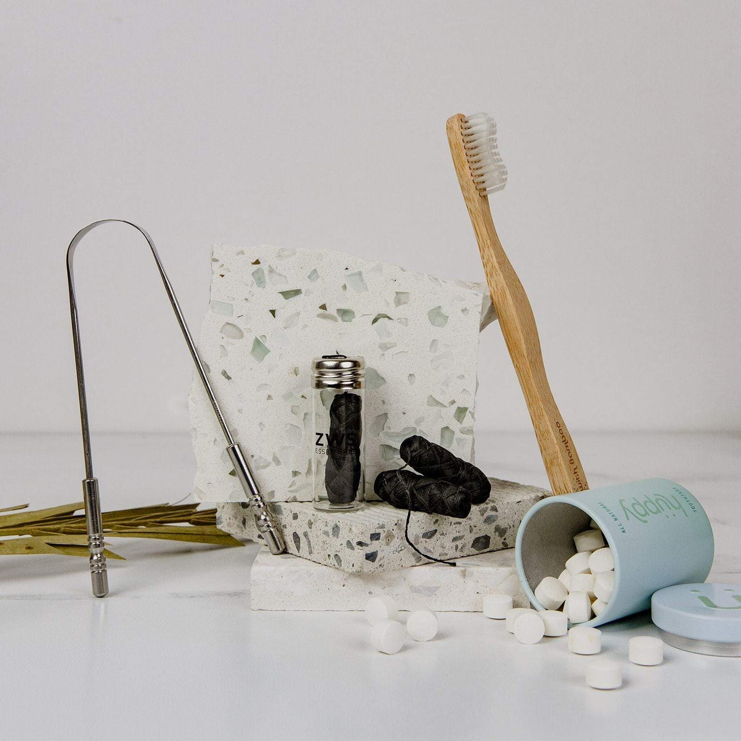 ZeroWasteStore.com’s Bamboo Charcoal Floss, plus a bamboo toothbrush, metal tongue scraper, and white toothpaste tablets are displayed together on a terrazzo-like surface for an eco-friendly dental routine.