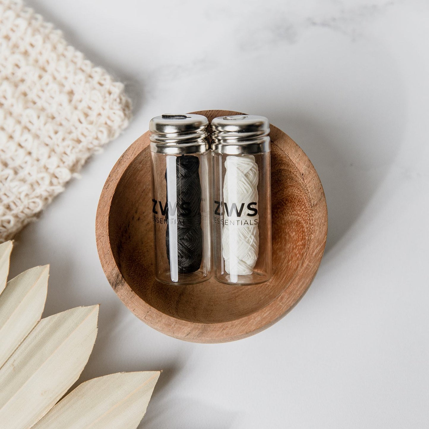 Two small glass jars with metal lids, each holding ZeroWasteStore.com Bamboo Charcoal Floss, sit in a wooden bowl on a white surface. Nearby are a knitted fabric and beige plant leaves.