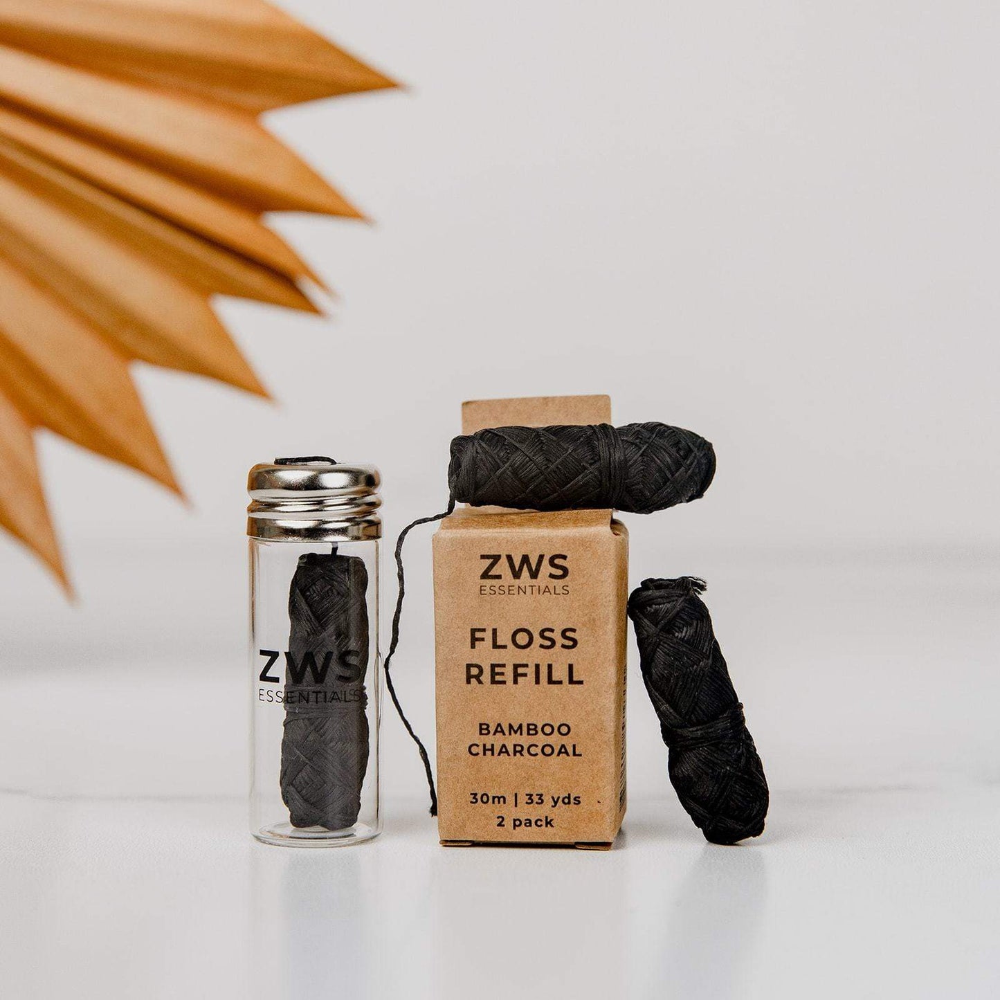 A glass ZeroWasteStore.com Bamboo Charcoal Floss jar sits beside its cardboard refill box, with two biodegradable spools of dental floss, displayed on a white surface.