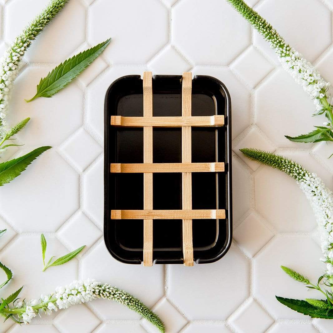 The Bamboo Soap Dish by ZeroWasteStore.com, featuring a black rectangular design with a wooden lattice cover, rests on white hexagonal tiles accented by green leaves and white flowering stems.