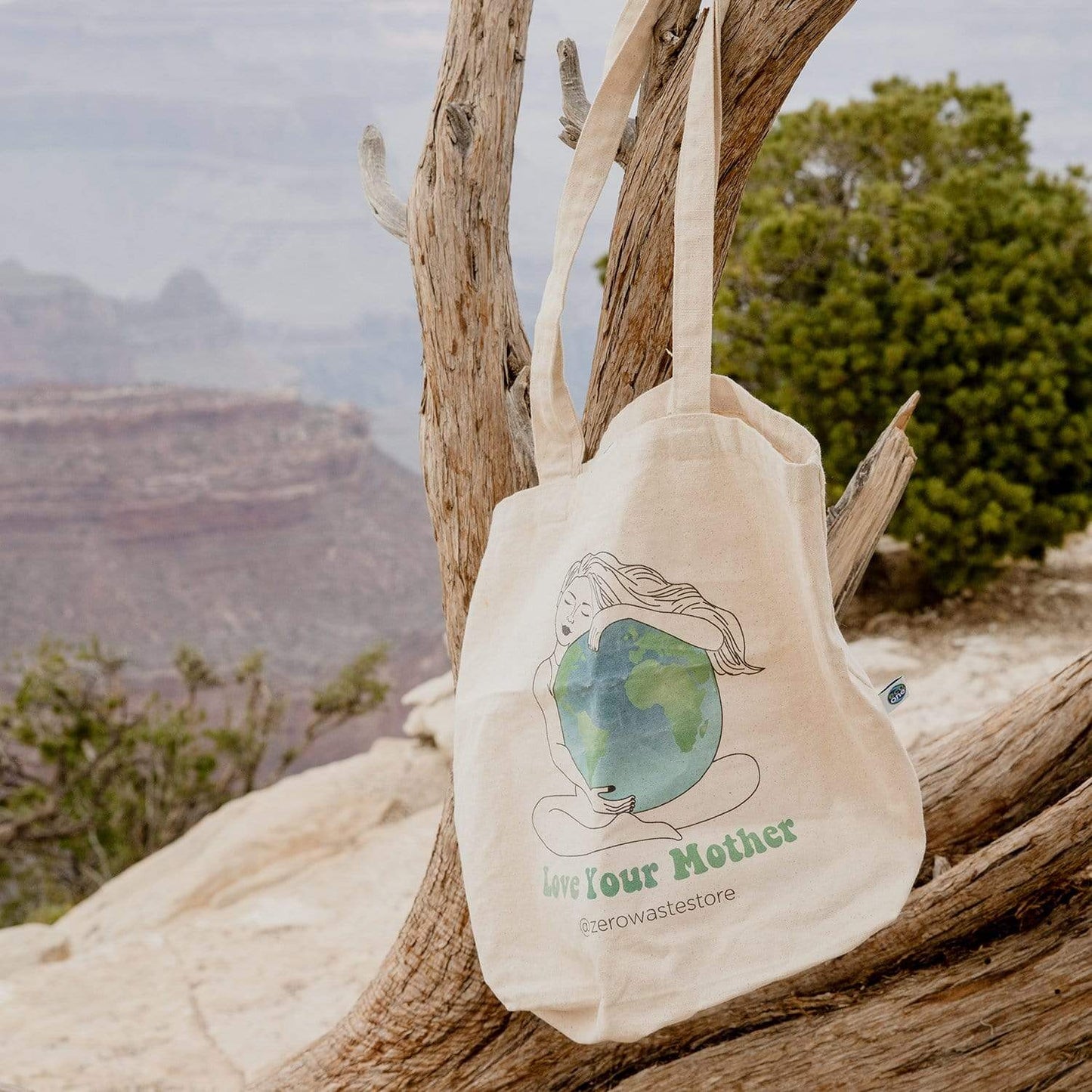 The Love Your Mother Organic Tote Bag from ZeroWasteStore.com, with an illustration of a woman hugging Earth, hangs on a twisted tree branch against a scenic canyon backdrop.