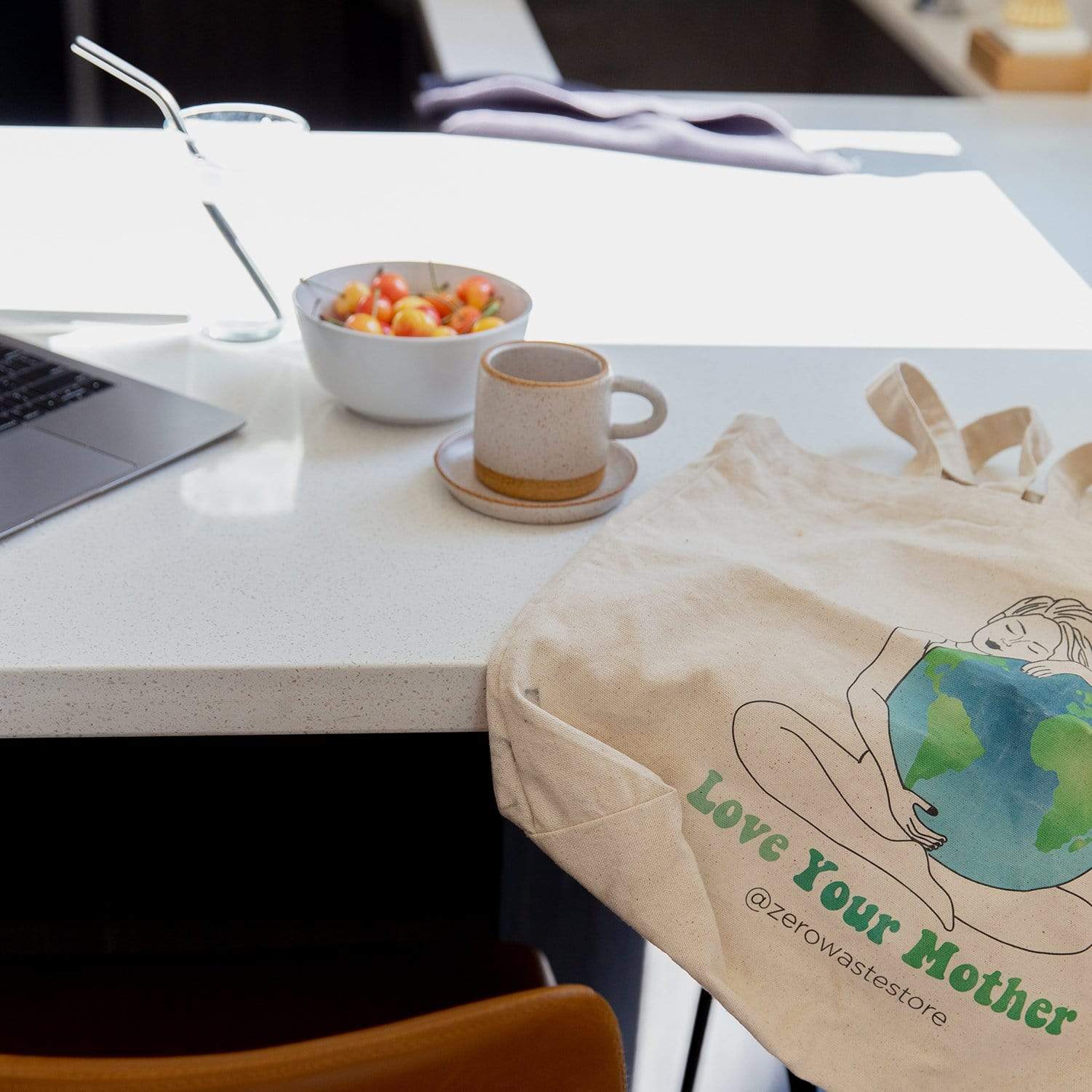The Love Your Mother Organic Tote Bag by ZeroWasteStore.com, featuring a woman hugging Earth and the words "Love Your Mother," sits on a kitchen counter next to a laptop, cup, fruit bowl, and glass of water.