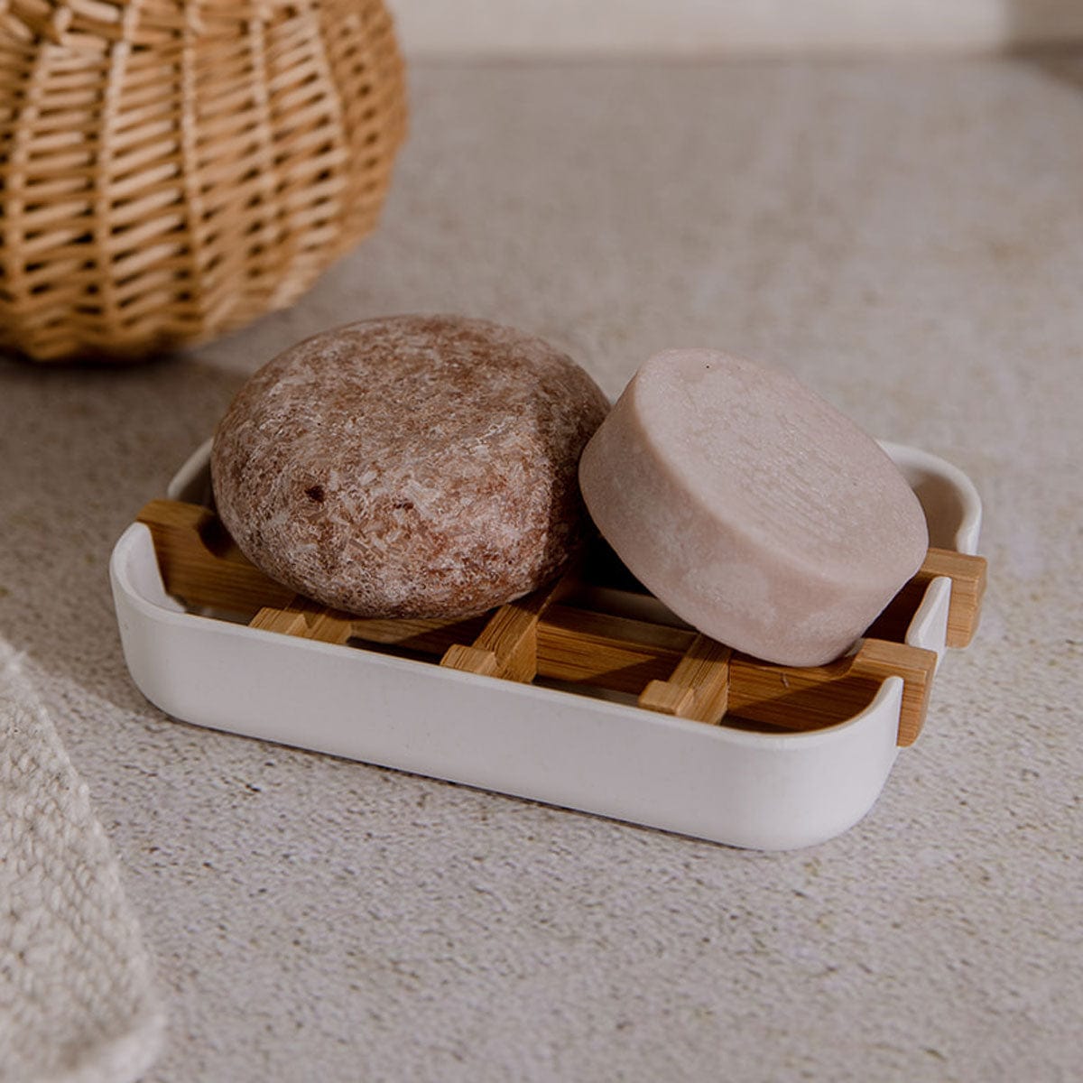 Two round bars of soap sit on the ZeroWasteStore.com Bamboo Soap Dish with a white tray, atop a speckled countertop. This plastic-free accessory complements a woven basket for an eco-friendly bathroom look.