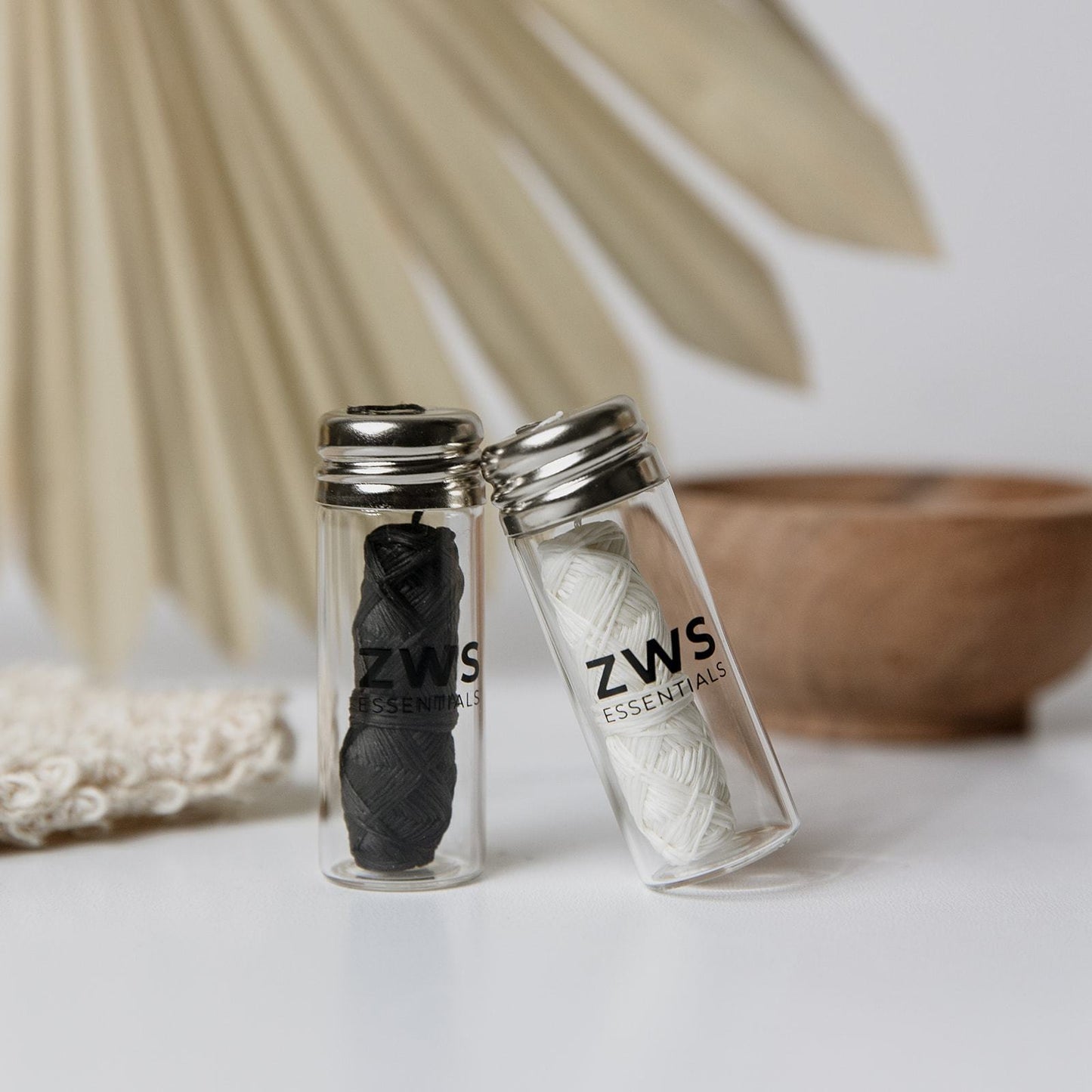 Two small glass jars labeled ZeroWasteStore.com contain Silk Floss – Zero Waste Dental Floss (30m, biodegradable and refillable) on a white surface with a blurred dried palm leaf and wooden bowl in the background.