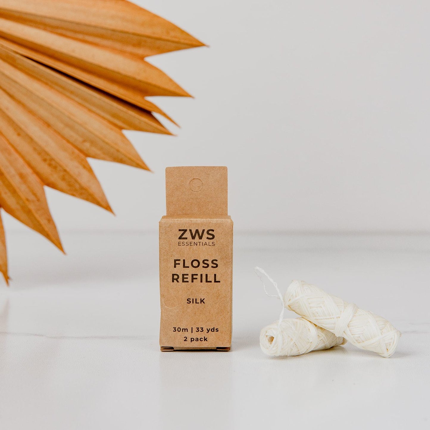 A ZeroWasteStore.com Silk Floss - Zero Waste Dental Floss, 30m, Biodegradable, Refillable box stands upright beside two silk floss rolls. A dried palm leaf appears in the background against a white surface.