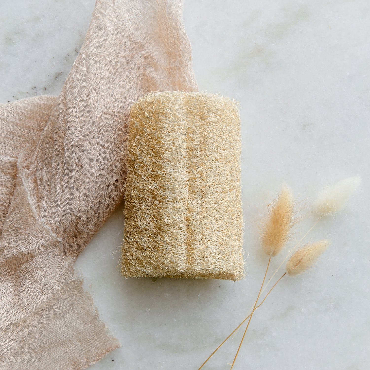 The Loofah Sponge Pack from ZeroWasteStore.com rests on light marble next to a soft beige cloth and dried flowers—an elegant, plastic-free choice for your zero waste routine.