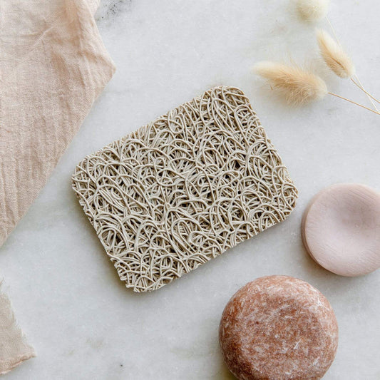 The ZeroWasteStore.com Soap Rest - Bar Soap Holder sits on marble beside a light tan cloth, a round brown bar of essential oils soap, a round pink dish, and dried wheat stems.