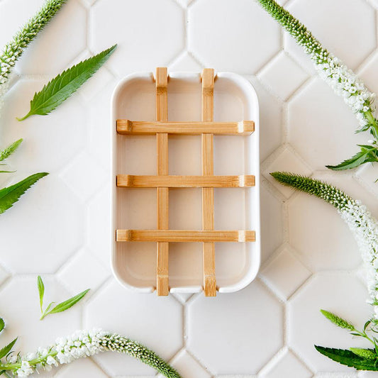 The Bamboo Soap Dish from ZeroWasteStore.com, featuring a white lattice design, rests on a white hexagonal tile surface with decorative green leaves and white flowers nearby.