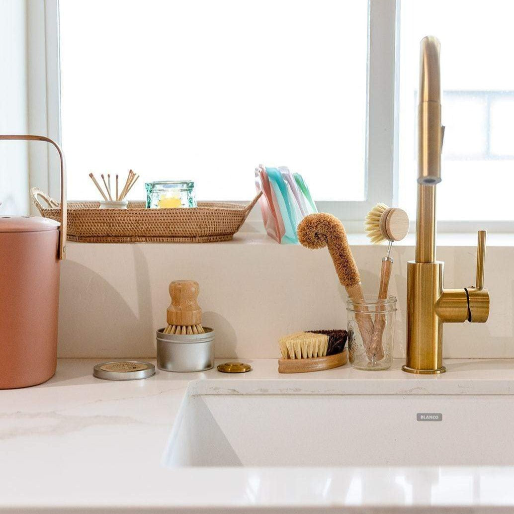 A ZeroWasteStore.com Zero Waste Dish Brush Kit with compostable brushes in a jar, plus a gold faucet, pink trash bin, soap dish, and wicker tray with candle and reed diffuser on a white countertop.