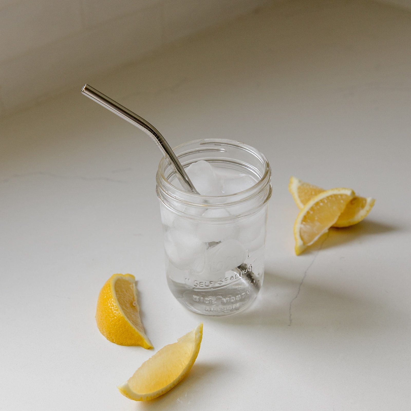 A mason jar of ice water with a ZeroWasteStore.com Stainless Steel Straw (Straight, Bent, or Boba Style) rests on a white countertop, surrounded by lemon wedges for a refreshing, plastic-free drink.
