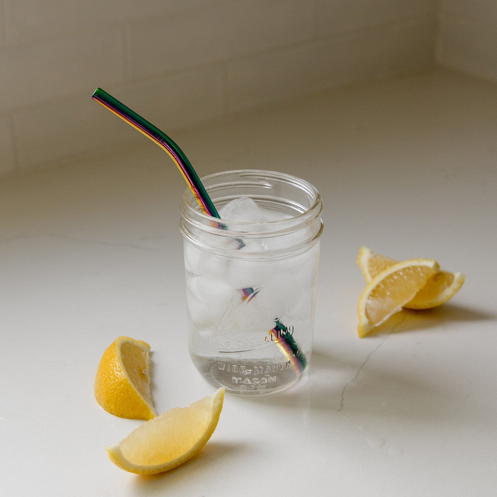A mason jar of ice water with a ZeroWasteStore.com Stainless Steel Straw (Straight, Bent, or Boba Style) rests on a white countertop beside three lemon wedges—a refreshing, plastic-free hydration choice.
