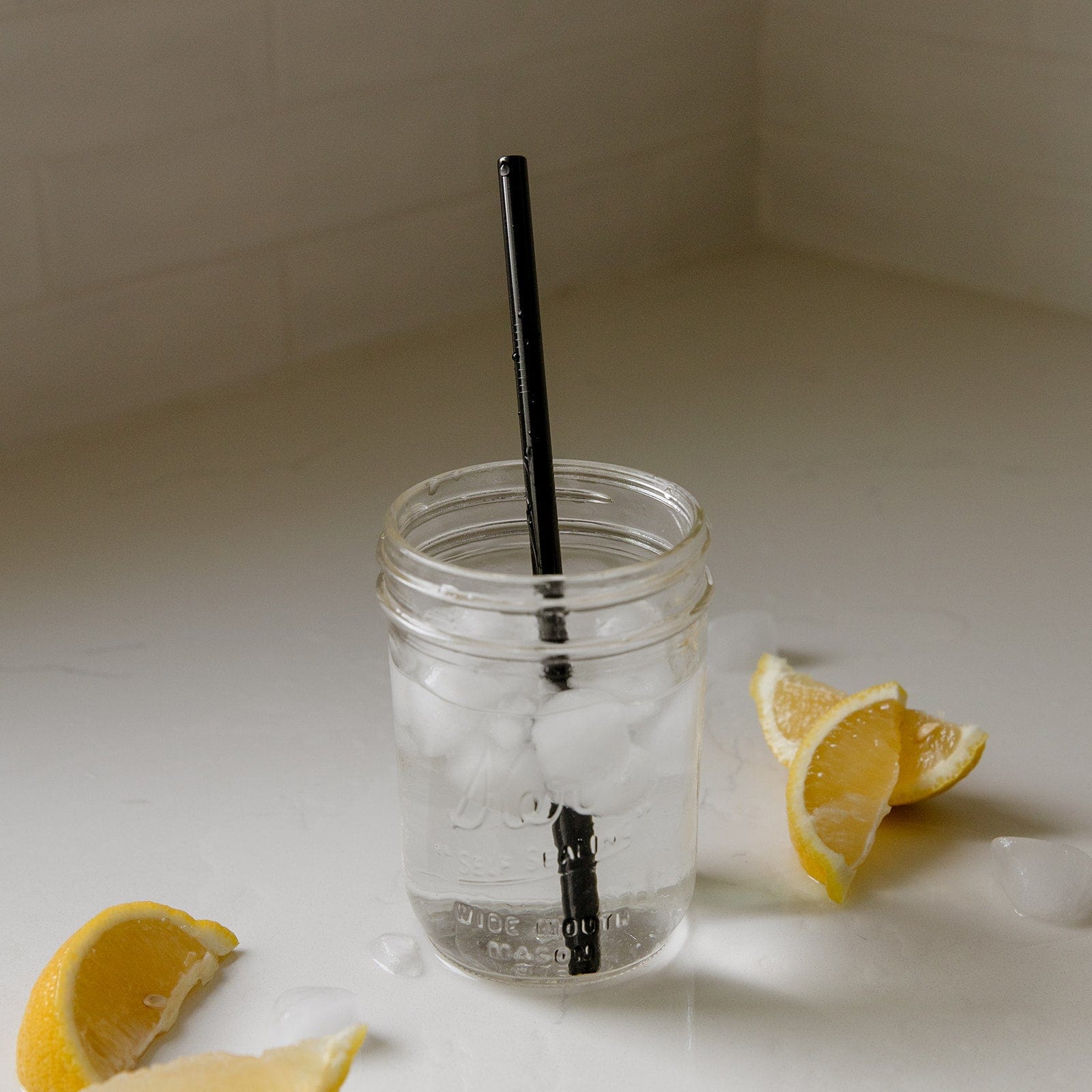 A mason jar with ice water and a ZeroWasteStore.com Stainless Steel Straw - Straight, Bent, or Boba Style sits on a white countertop, surrounded by ice cubes and lemon wedges.