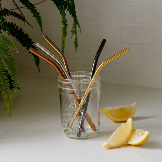 A mason jar on a white counter holds five colorful Stainless Steel Straws (Straight, Bent, or Boba Style) from ZeroWasteStore.com, beside lemon wedges; fern leaves and a white tiled wall create a bright, plastic-free setting.