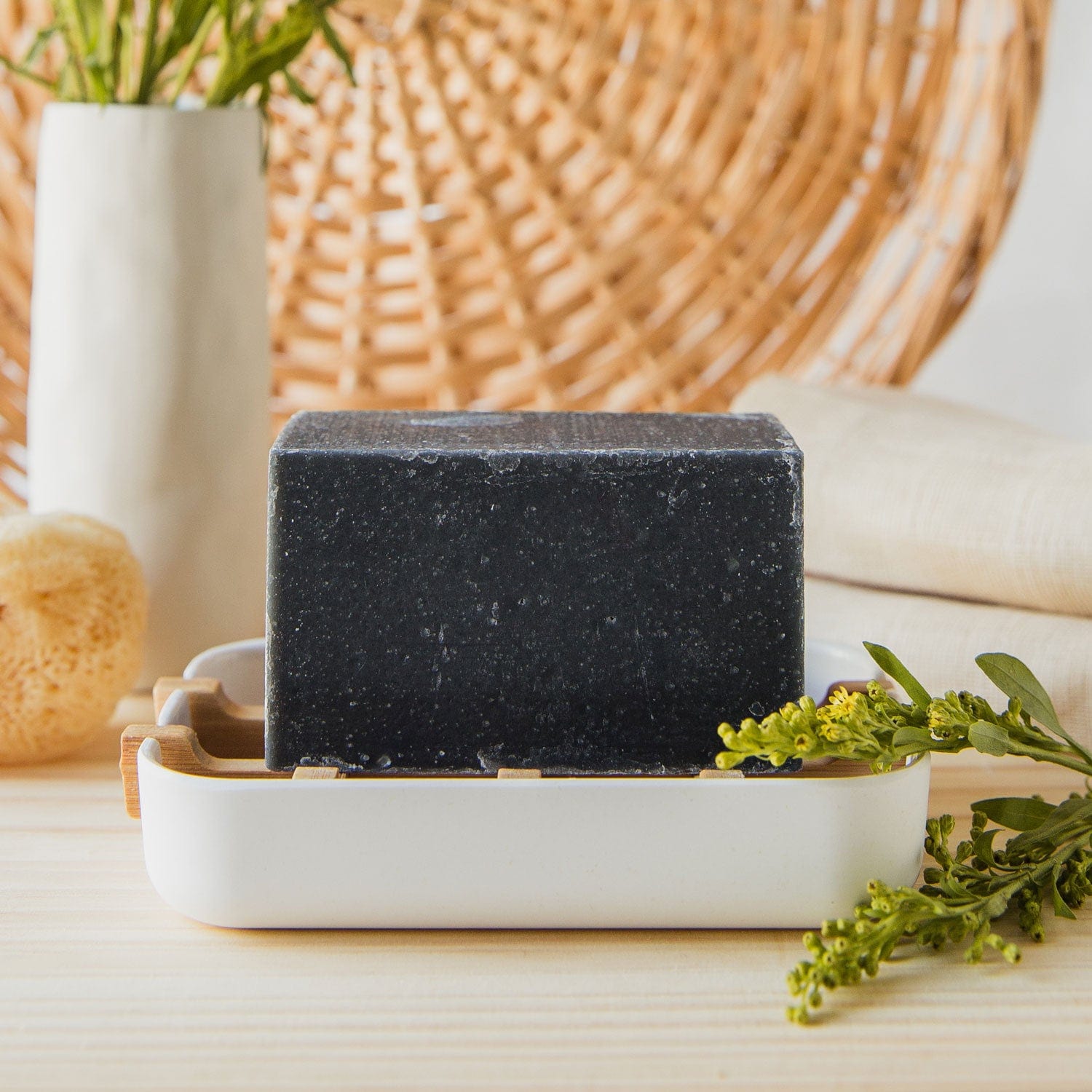 A rectangular black 4oz all natural, vegan bar soap from ZeroWasteStore.com sits on a soap dish beside yellow flowers, a sponge, towel, and vase. The zero waste soap comes in 6 scents and is plastic free—perfect for face and body care.