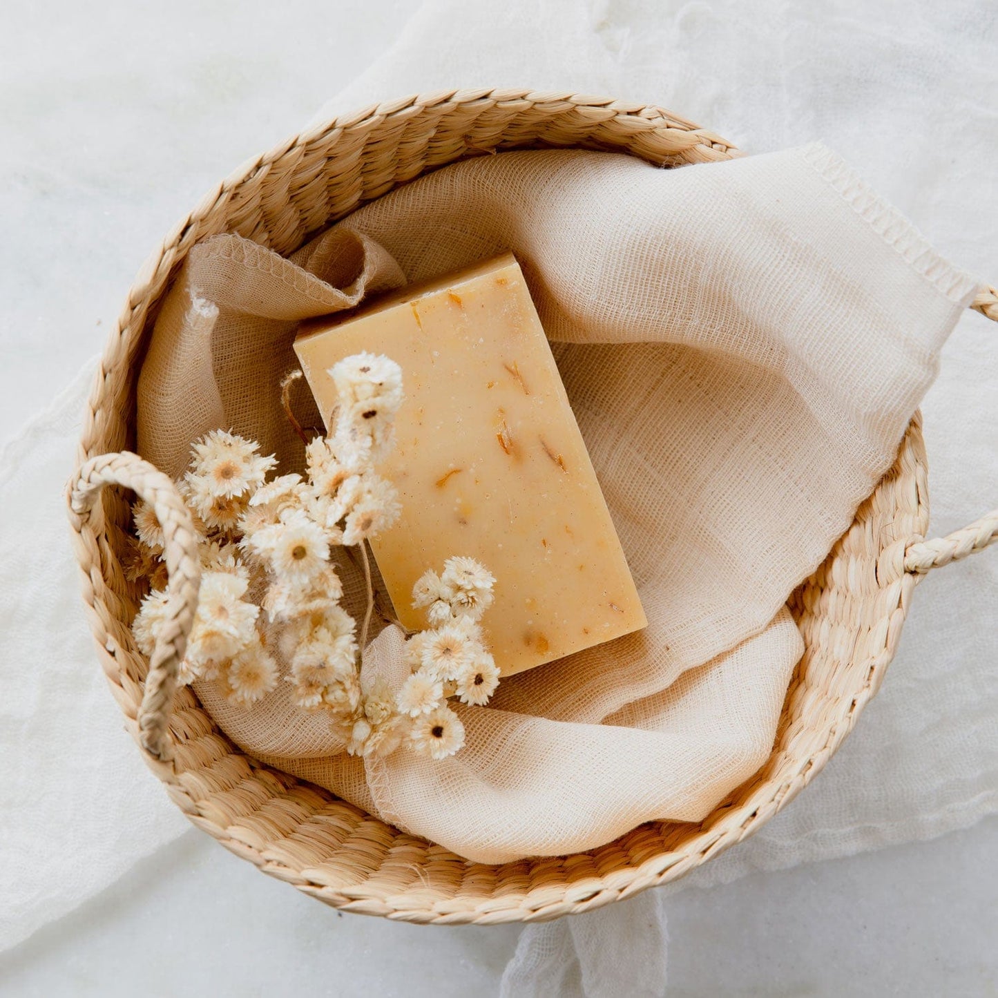 A 4oz all natural, vegan Soap Bar from ZeroWasteStore.com rests on a beige cloth in a woven basket, surrounded by dried white flowers. Available in 6 scent options; plastic free and suitable for face and body.
