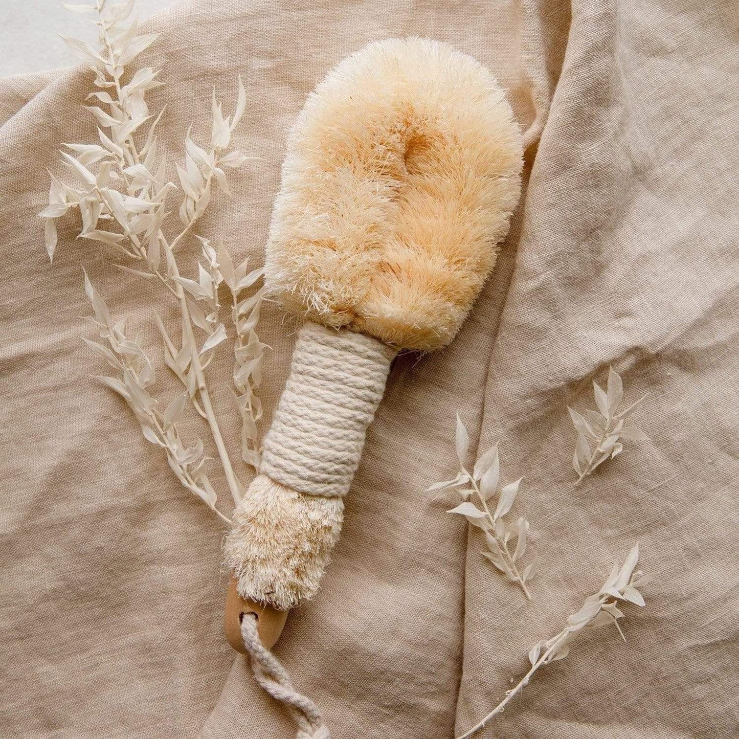 The ZeroWasteStore.com Dry Body Brush features sisal bristles and a rope handle, shown on beige linen with dried white leaves—ideal for gentle exfoliation and lymphatic drainage.