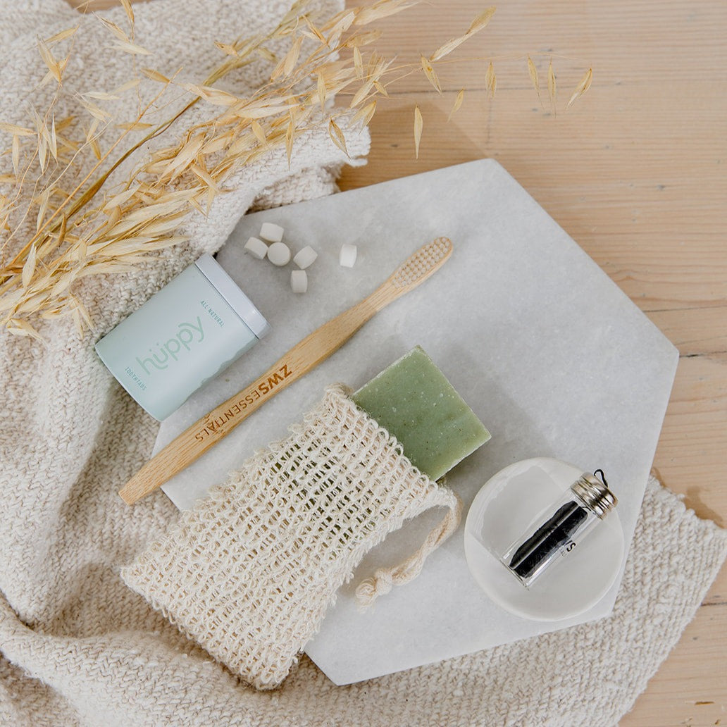 A flat lay of zero waste bathroom essentials, featuring a bamboo toothbrush, toothpaste tablets, a green bar of soap in the ZeroWasteStore.com Soap Saver Bag, a small glass vial, and dried wheat on a white hexagonal tray.