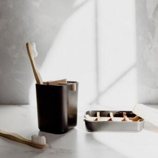 The Zero Waste Brush Holder & Soap Dish Bundle from ZeroWasteStore.com features bamboo toothbrushes in a black holder, a compostable brush on a white surface, and an open stainless steel soap dish—sunlit shadows create an eco-friendly bathroom scene.
