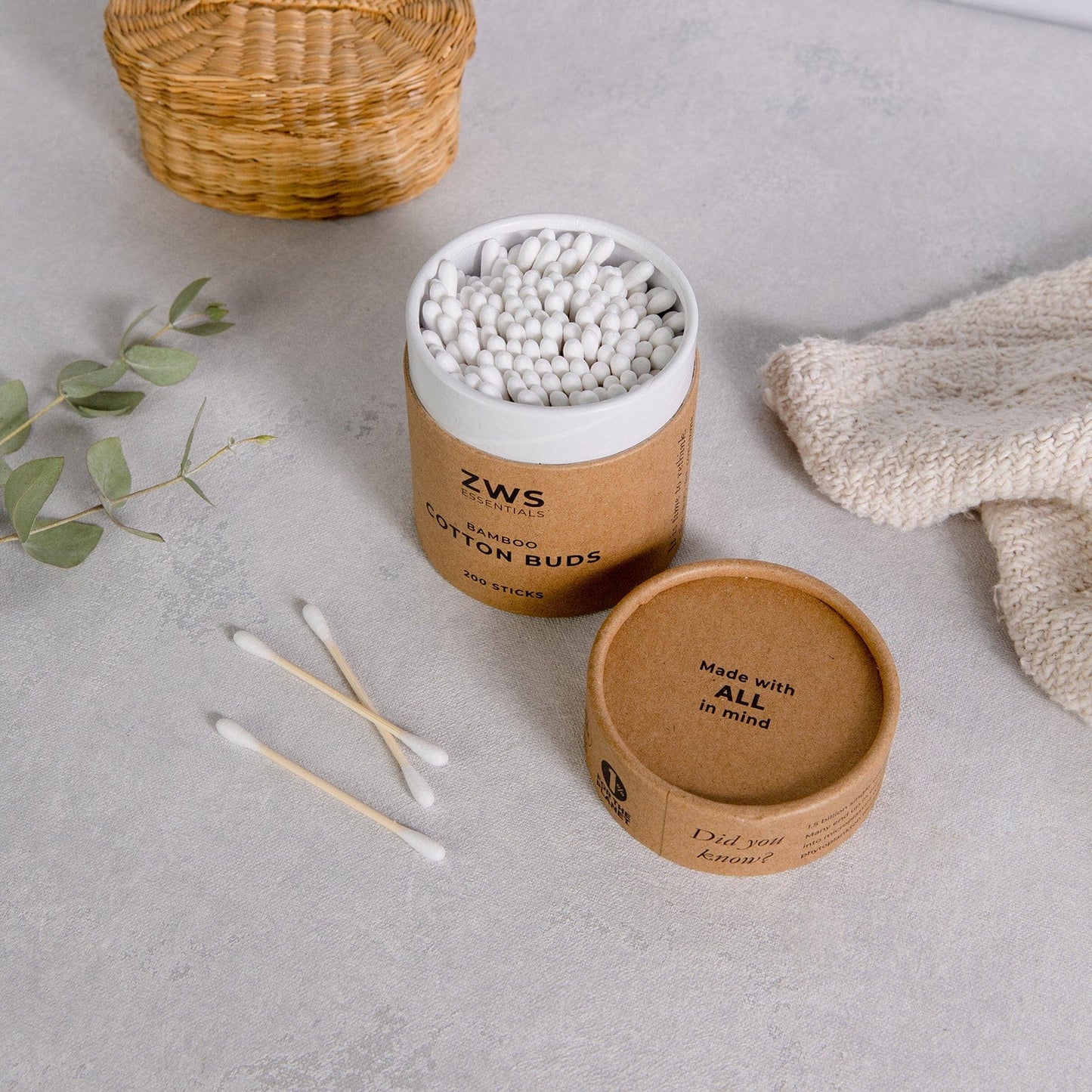 A container of ZeroWasteStore.com Bamboo Cotton Buds with the lid off rests on a light surface, three buds beside it. In the background are a wicker basket, a beige knitted cloth, and a sprig of greenery.