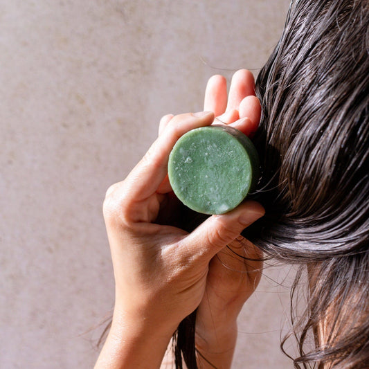 A person rubs a ZeroWasteStore.com Conditioner Bar onto their wet, dark hair with both hands visible against a neutral, light background.
