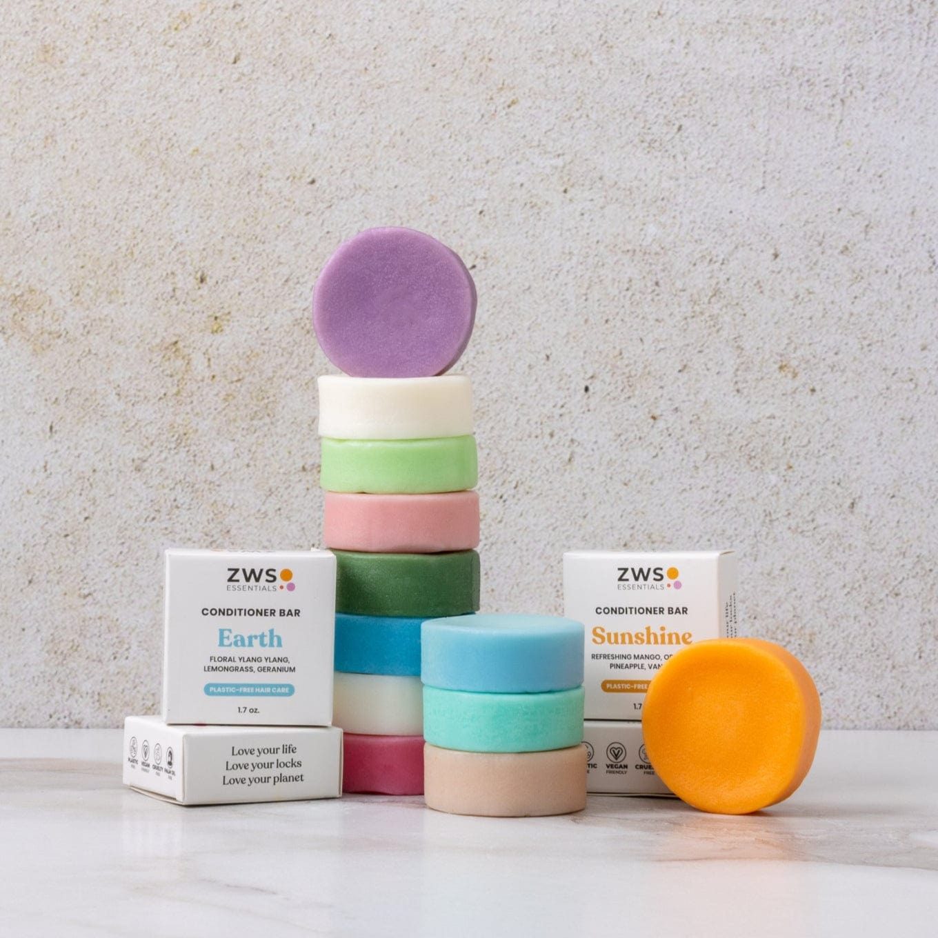 A stack of colorful ZeroWasteStore.com Conditioner Bars—including a vegan, sulfate-free option—stands between Earth and Sunshine boxes. An orange bar rests against the Sunshine box on a neutral stone background.