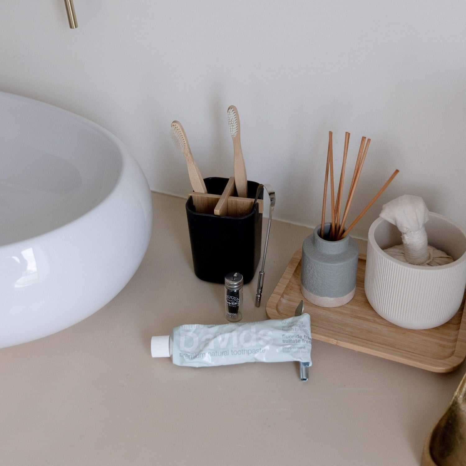 On the bathroom counter: a sink, a black cup with two bamboo toothbrushes and metal tweezers, a small jar, ZeroWasteStore.com Bamboo Charcoal Floss, a gray reed diffuser container, a tissue tray, and toothpaste.