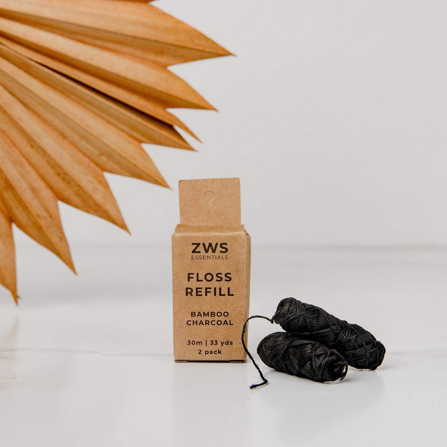 A box labeled ZeroWasteStore.com Bamboo Charcoal Floss sits beside two rolls of biodegradable dental floss on a white surface, with a dried palm leaf in the background.