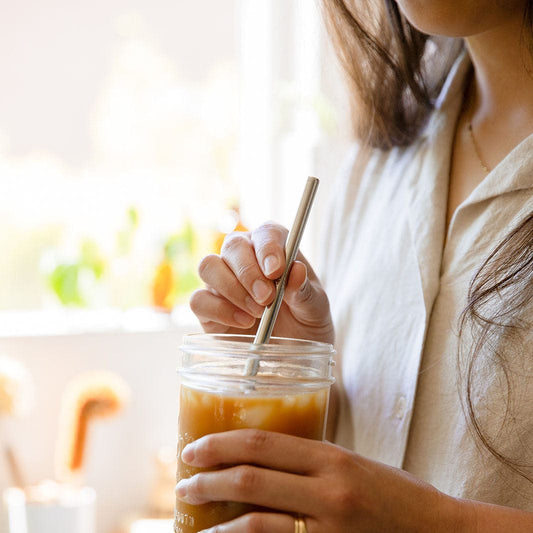 A person stirs an iced drink with a ZeroWasteStore.com Stainless Steel Straw—Straight, Bent, or Boba Style—in a glass jar near a sunny window, choosing a plastic-free lifestyle.