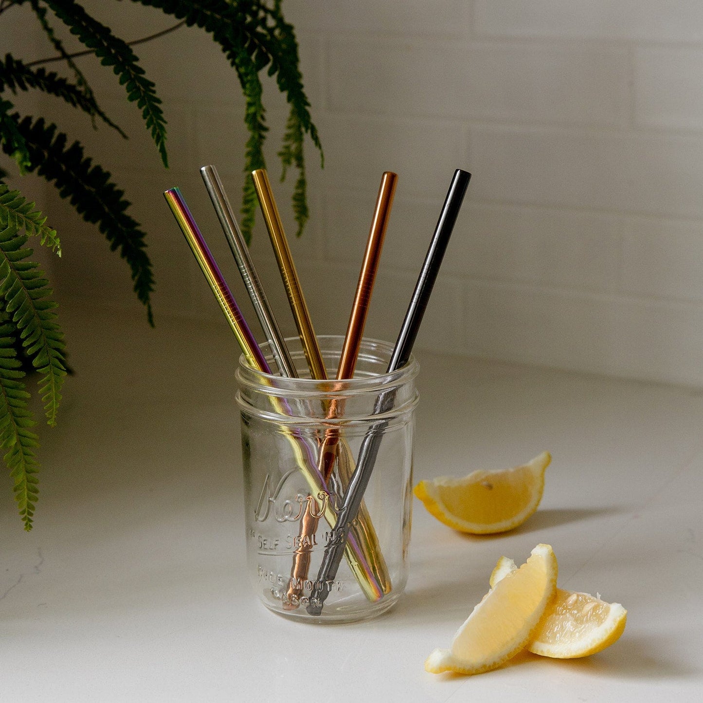 A mason jar with five ZeroWasteStore.com Stainless Steel Straws (straight, bent, or boba style) in assorted metallic colors sits on a white countertop, accented by lemon wedges and fern leaves for a chic plastic-free display.