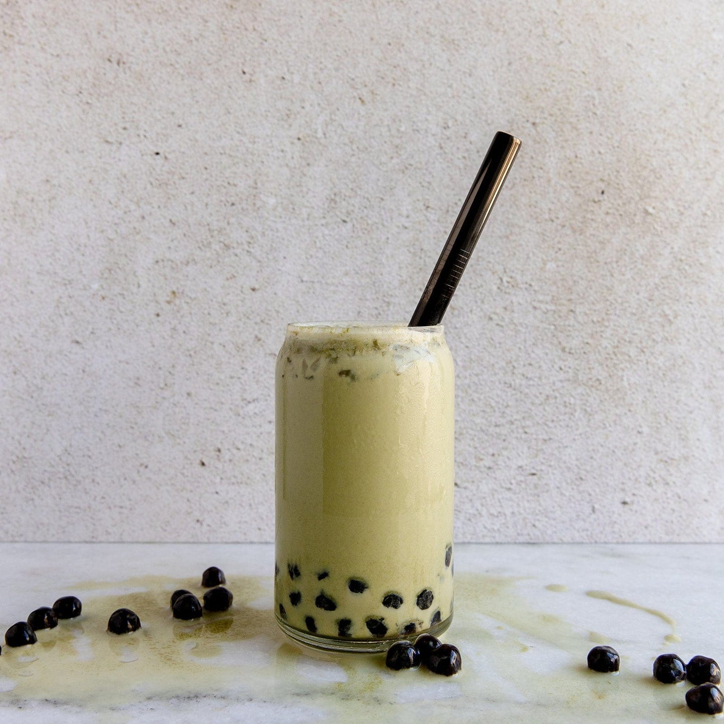 A glass of matcha bubble tea with black tapioca pearls features the ZeroWasteStore.com Stainless Steel Straw - Straight, Bent, or Boba Style. Some tea and pearls are spilled on a white marble surface; the backdrop is a plain light-colored wall.