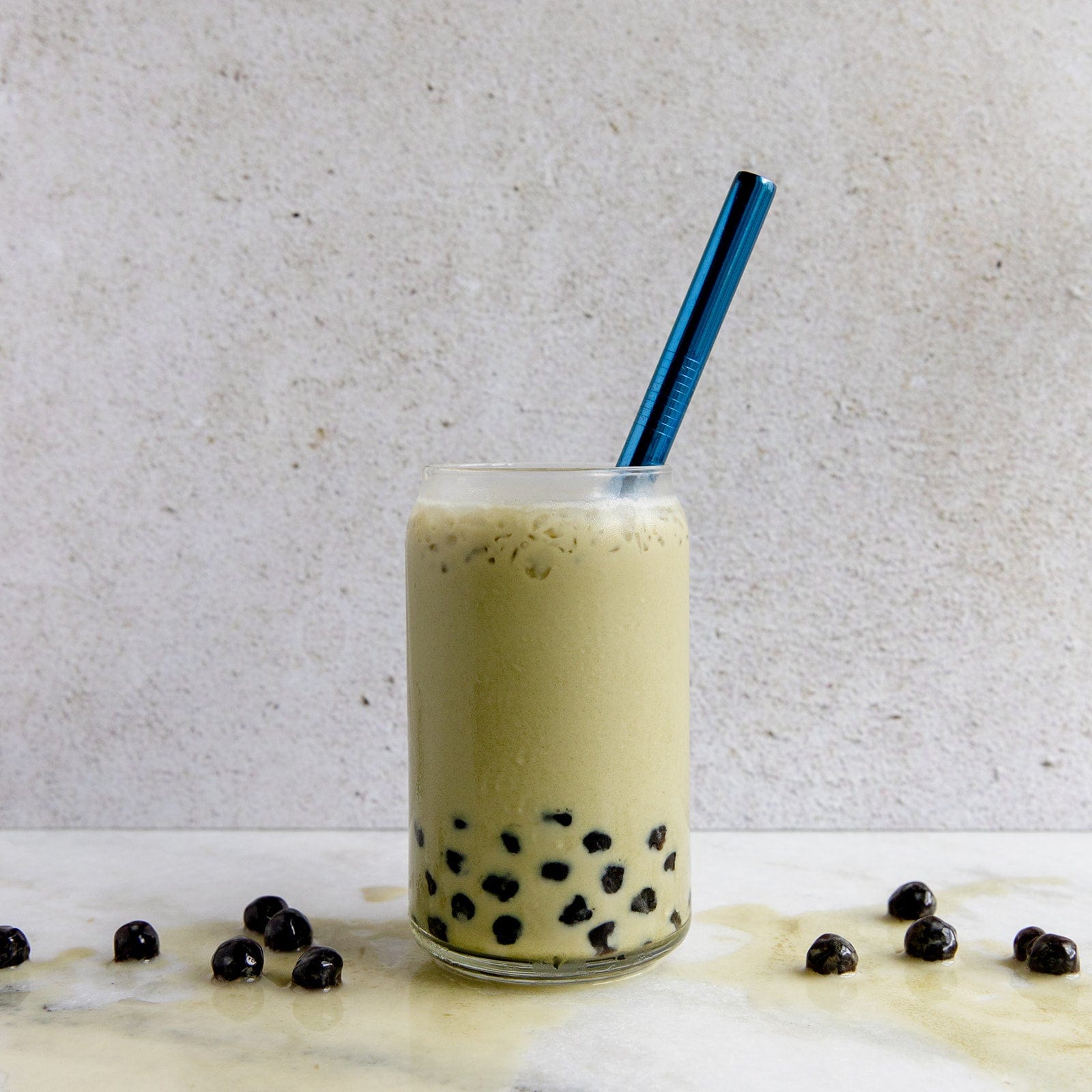 A glass of creamy light brown milk tea with black tapioca pearls sits on a white surface, featuring a ZeroWasteStore.com Stainless Steel Straw - Straight, Bent, or Boba Style, with scattered pearls around it.