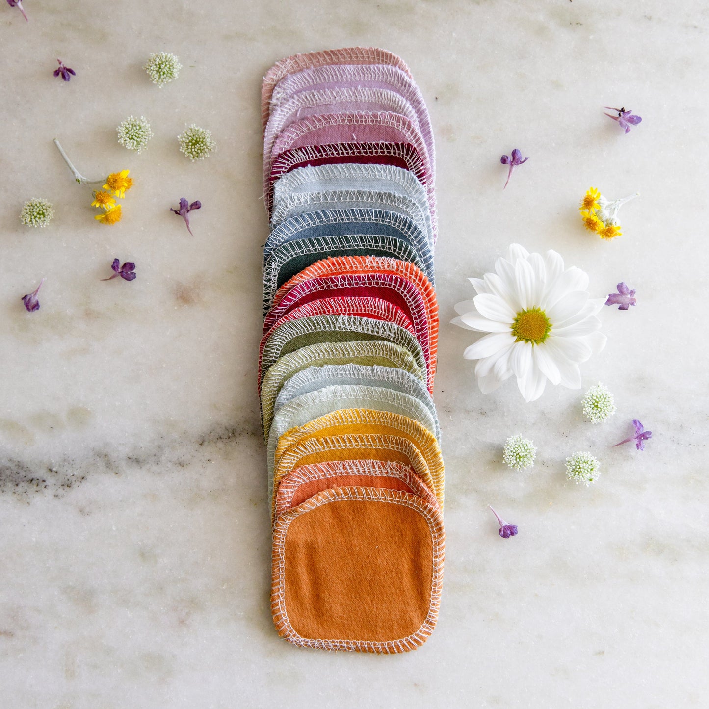 ZeroWasteStore.com Reusable Cotton Rounds are neatly stacked on a marble surface, surrounded by small white, yellow, and purple flowers, with a single white daisy placed to the right.