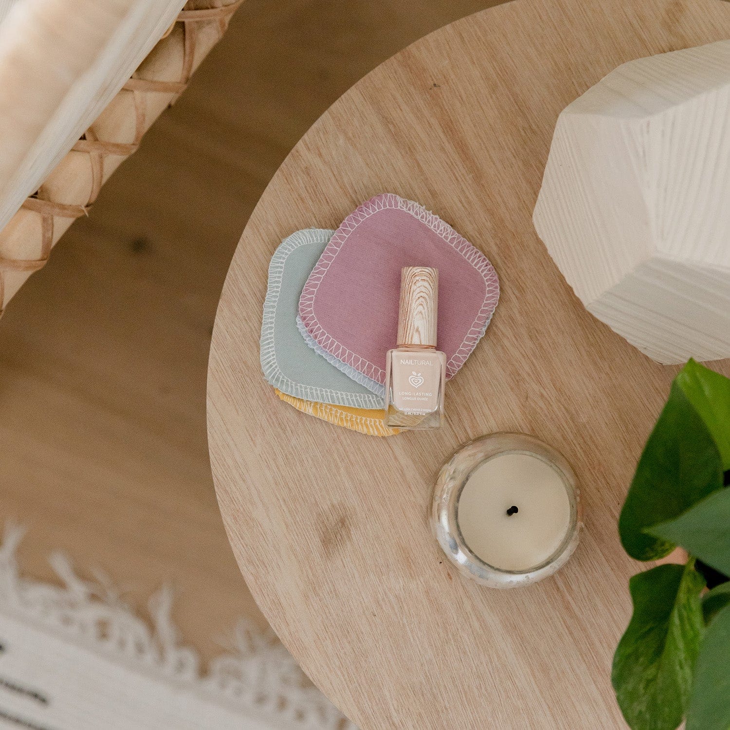 On a wooden table: a nail polish bottle, three pastel Reusable Cotton Rounds by ZeroWasteStore.com, a lit candle in a glass holder, and a green plant in the corner.