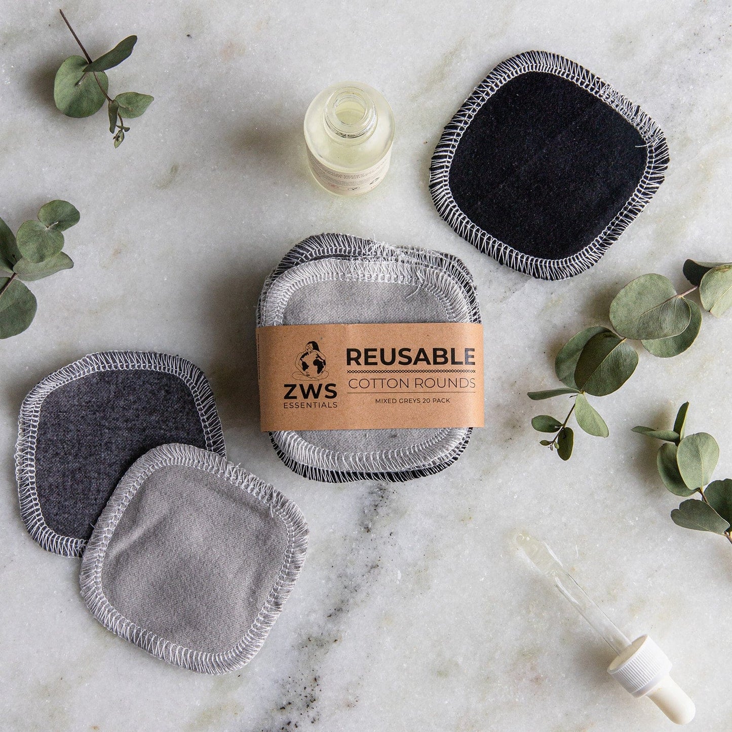 A stack of Reusable Cotton Rounds by ZeroWasteStore.com in gray and black, with a brown paper label, sits on marble beside eucalyptus leaves and a dropper—ideal for sustainable skincare routines.