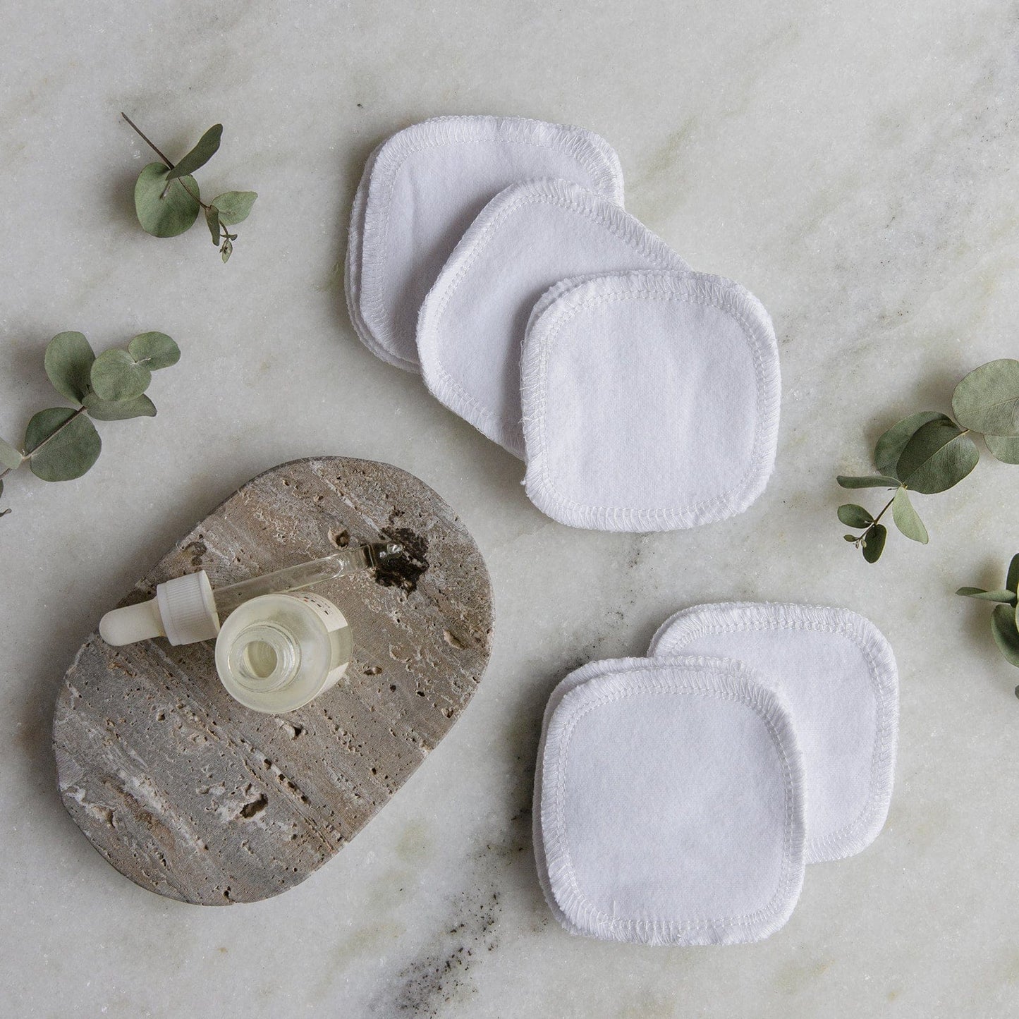 ZeroWasteStore.com's Reusable Cotton Rounds are displayed on a marble surface with a dropper bottle on a stone coaster, surrounded by eucalyptus sprigs for an eco-friendly, calming vibe.