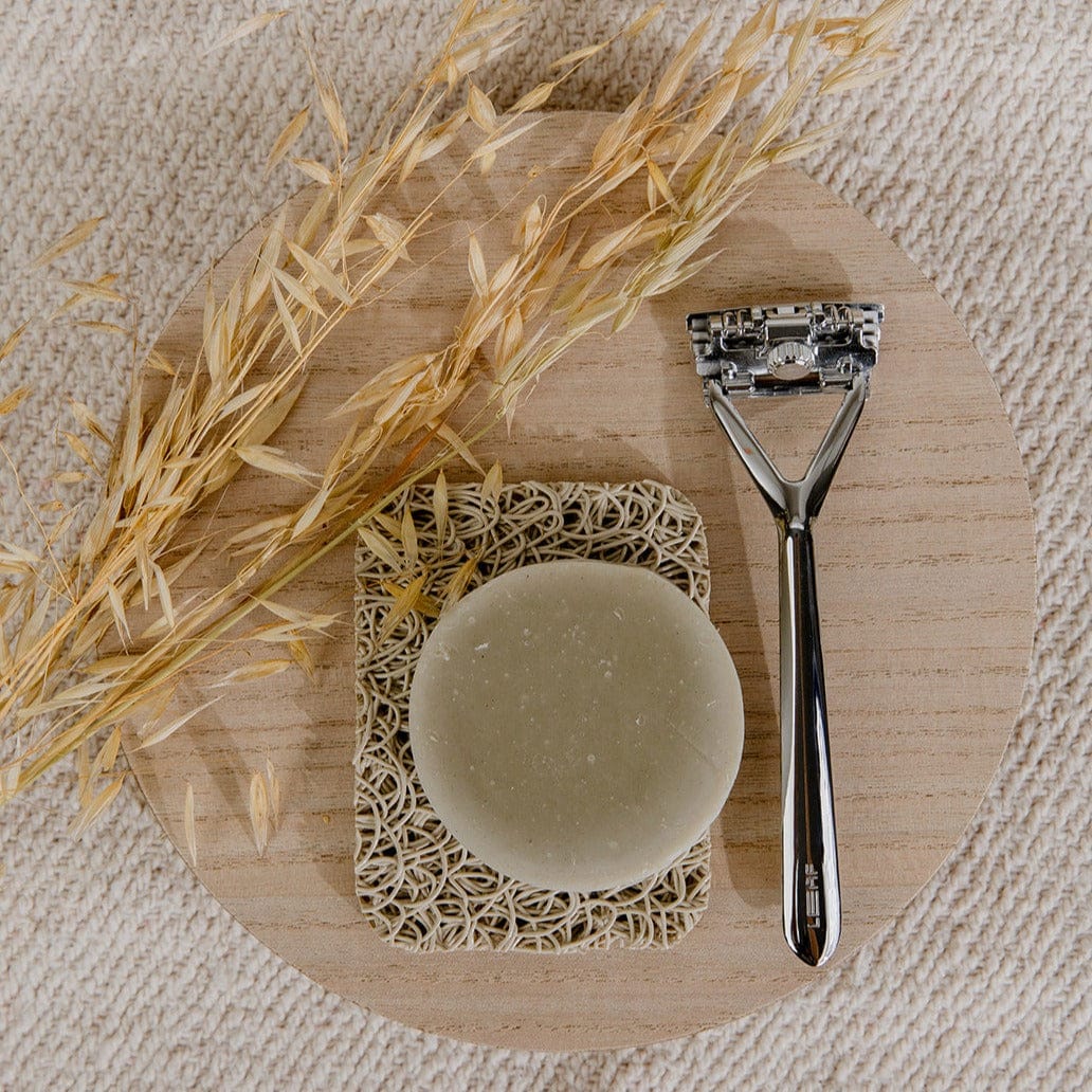 A bar of soap rests on the ZeroWasteStore.com Soap Rest—Bar Soap Holder, Bar Soap Lift—beside a metal razor and dried oats, all arranged on a round wooden tray atop a textured beige surface.