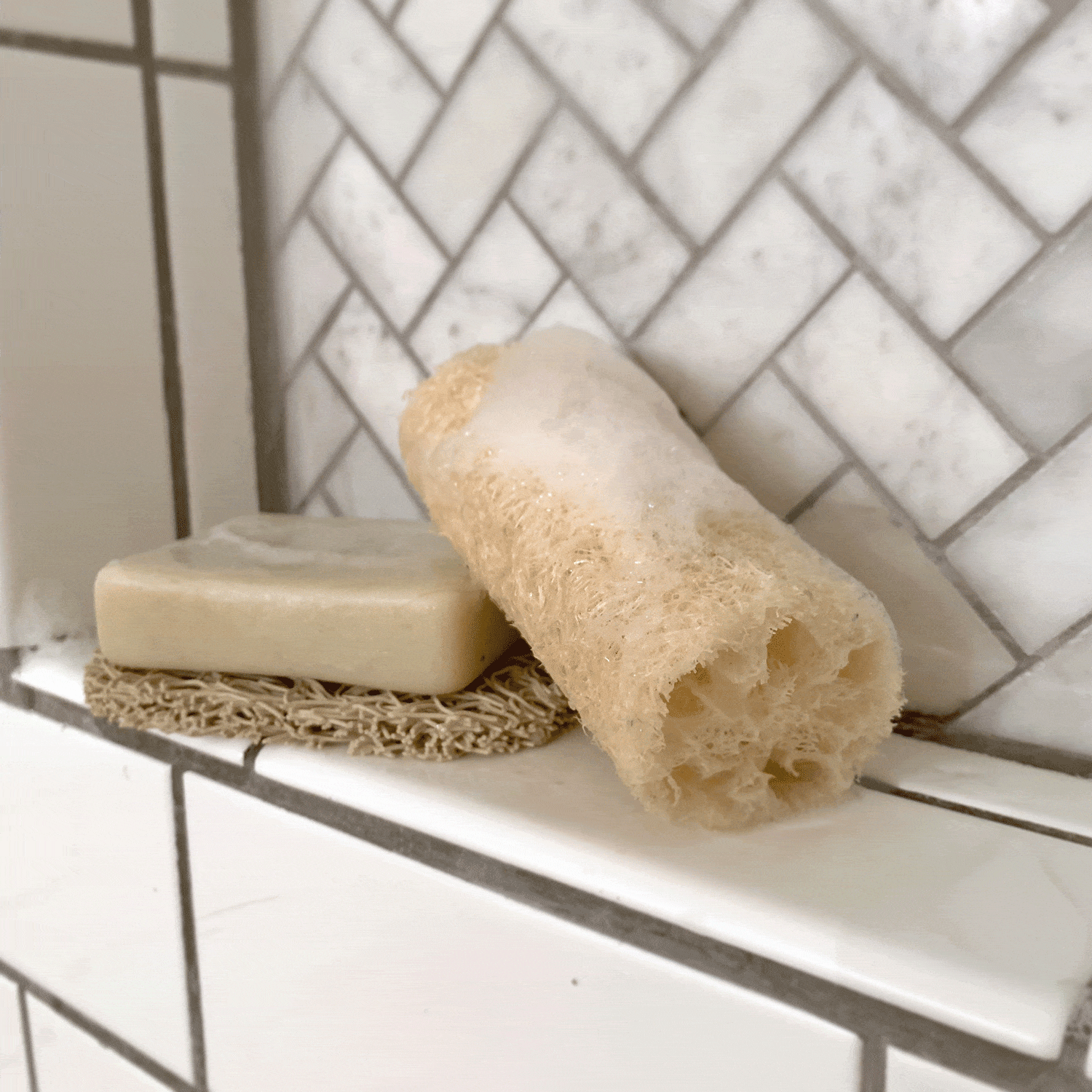 A beige bar soap sits on the ZeroWasteStore.com Soap Rest—Soap Holder, Bar Soap Rest, Lift for Bar Soap—with a natural loofah and suds on a tiled shower shelf against a white herringbone backsplash.