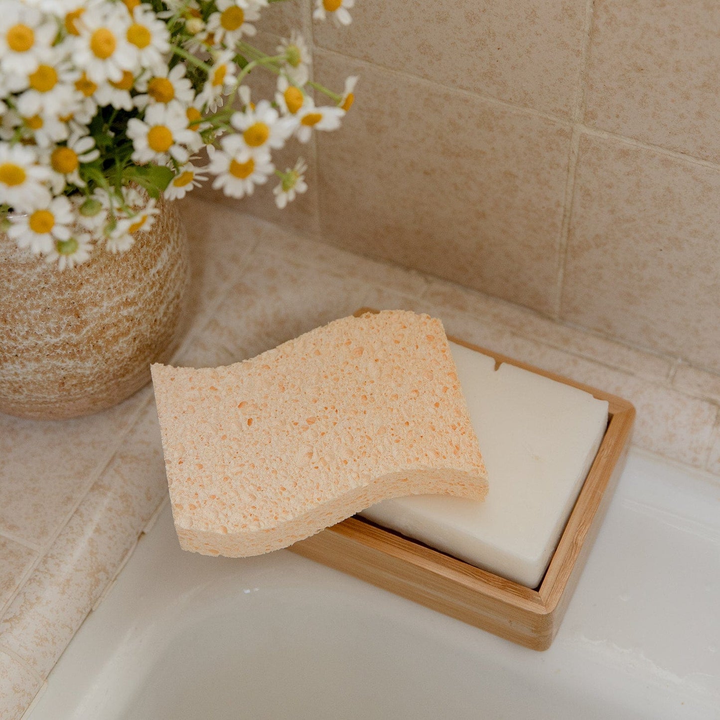 A ZeroWasteStore.com Cellulose Kitchen Sponge sits on a white soap bar in a wooden dish by the sink, with daisies in a vase nearby on the tiled bathroom countertop.