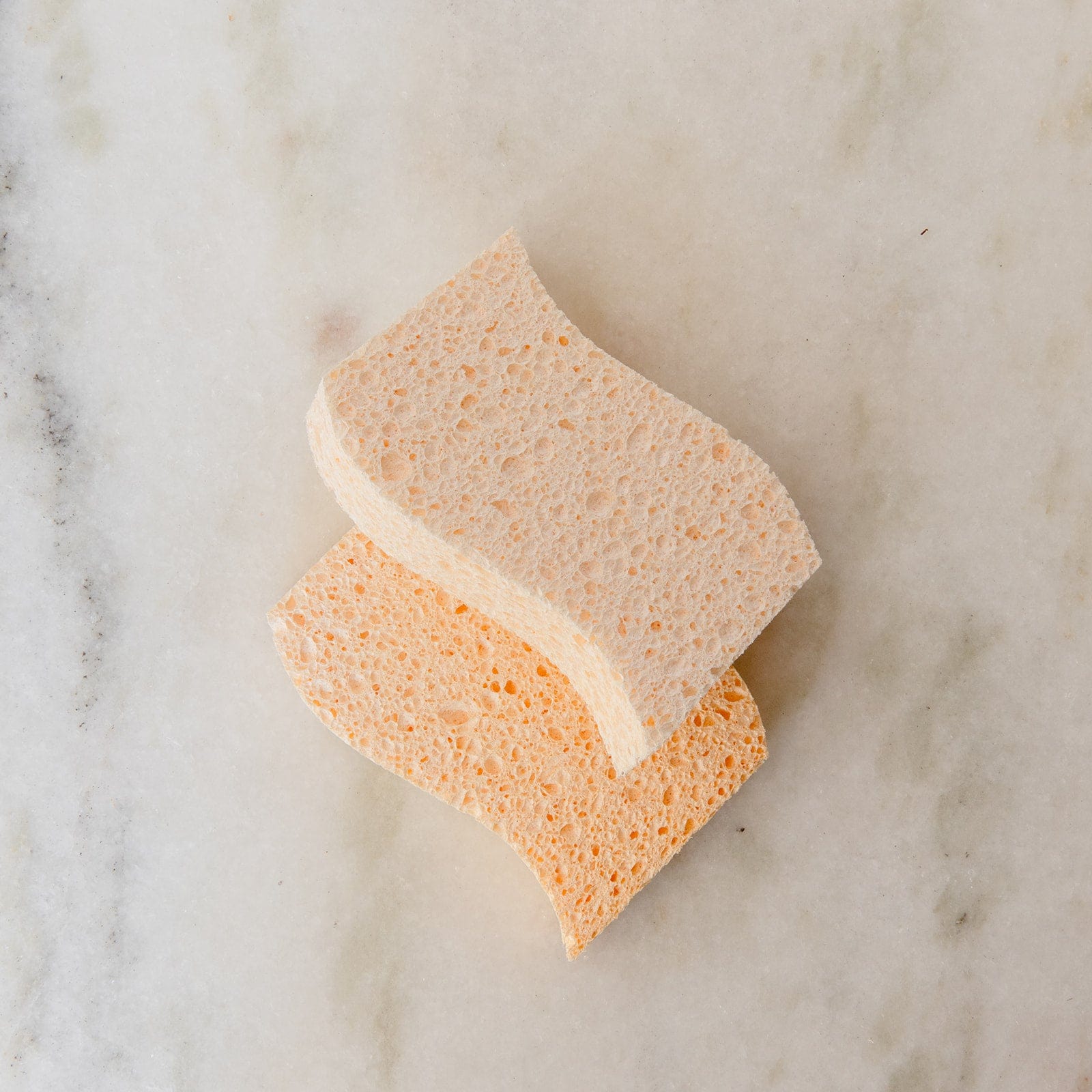Two rectangular Cellulose Kitchen Sponges from ZeroWasteStore.com, one beige and one light orange, are stacked on a marble-like surface. These biodegradable sponges have a wavy edge and porous texture for effective cleaning.